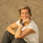 Beatriz Llamas Mentora Financiera sentada sobre la arena y sonriendo a la cámara. Vestida con vaquero gris y camiseta color crema