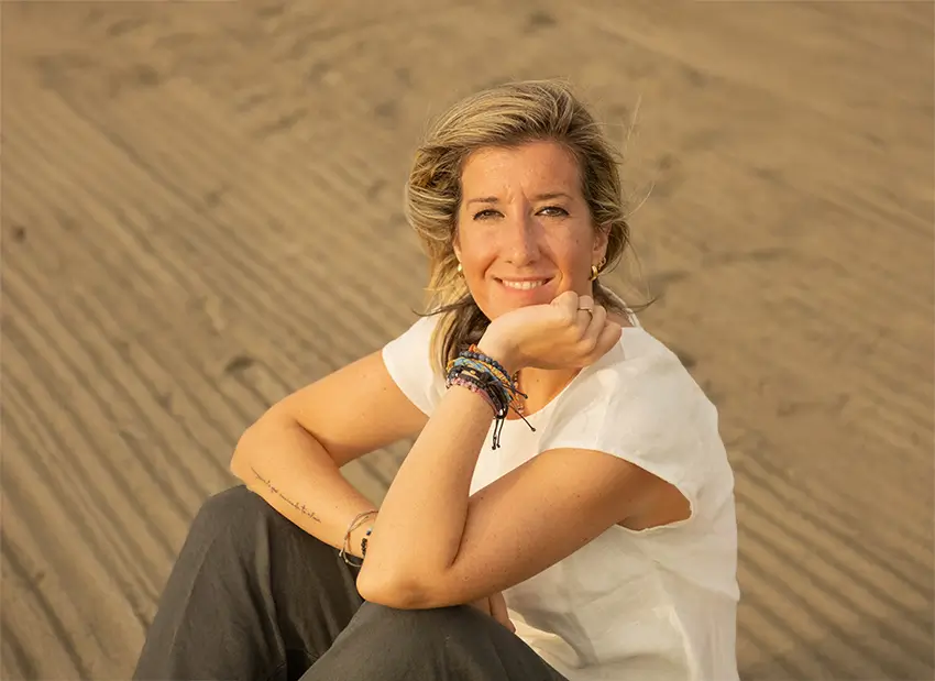 Beatriz Llamas Mentora Financiera sentada sobre la arena y sonriendo a la cámara. Vestida con vaquero gris y camiseta color crema