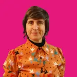 Virginia Romera en primer plano con fondo rosa mirando a la cámara con una camisa de colores